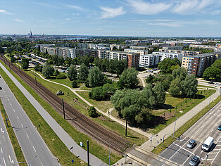 Luftaufnahme eines Wohngebiets mit mehrstöckigen Gebäuden, Bäumen, einem Gleisbett und einer Straße mit parkenden Autos