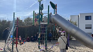 Kinder spielen auf einem Klettergerüst mit Rutsche auf einem Sandspielplatz, Erwachsene beobachten im Hintergrund