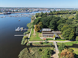 Luftaufnahme eines Flussufers mit Bootssteg, Gebäuden und angrenzendem Wald vor einer Stadt im Hintergrund