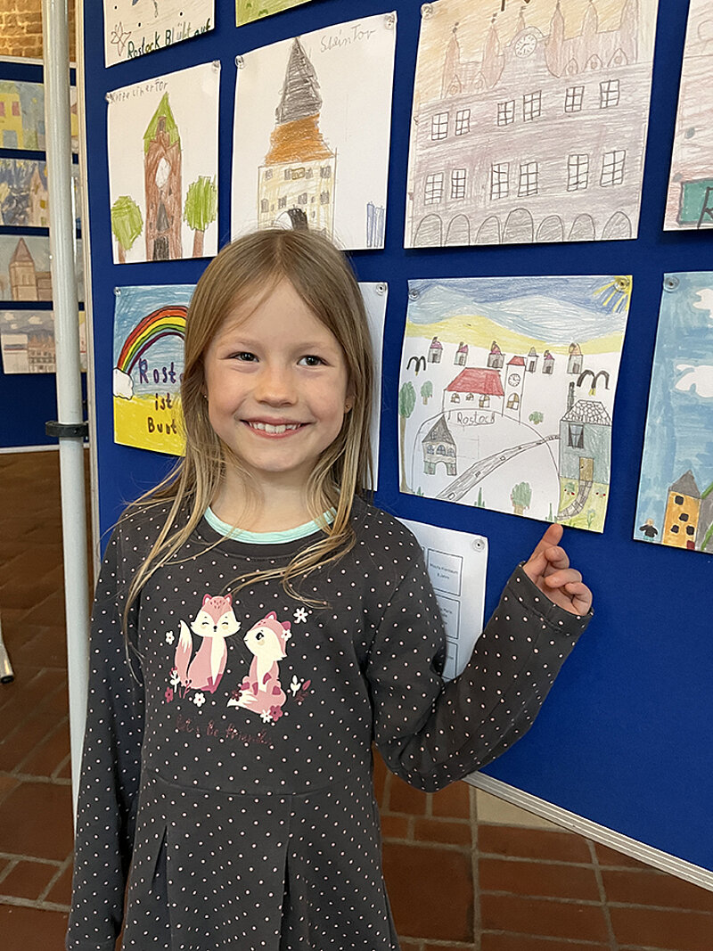Kind mit langem Haar und grauen Kleid zeigt ein Bild auf einer Wand mit mehreren Kinderzeichnungen mit Stadtansichten
