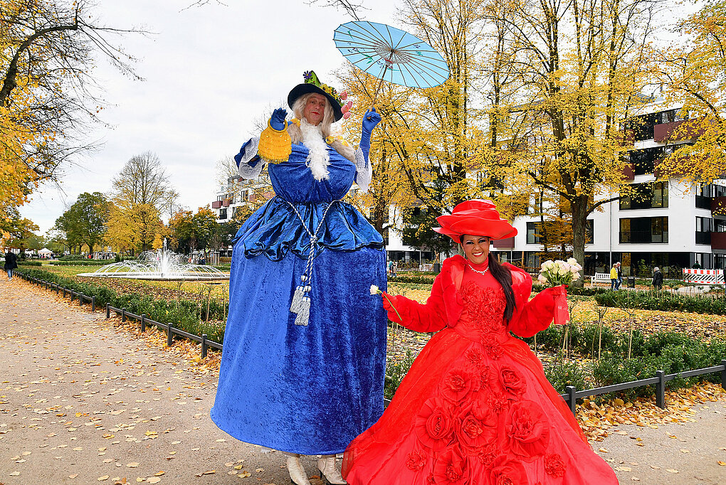 Zwei Personen in historischen Kostümen, eine in blauem und eine in rotem Kleid, vor dem Rosengarten bei sonnigem Wetter