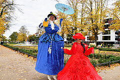 Zwei Personen in historischen Kostümen, eine in blauem und eine in rotem Kleid, vor dem Rosengarten bei sonnigem Wetter