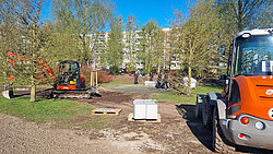 Baustelle in einem Park mit Bagger, Radlader und mehreren Personen im Hintergrund vor Wohngebäuden