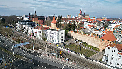 Luftaufnahme von Rostock mit historischen Gebäuden, darunter eine weiße Villa und eine Stadtmauer, umgeben von Straßen und Verkehr.