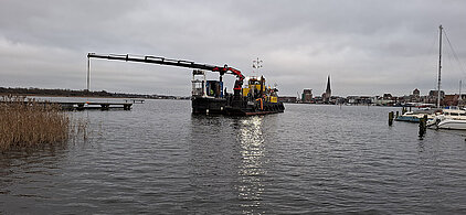 Arbeitsschiff mit Kran auf ruhigem Wasser unter bewölktem Himmel, im Hintergrund Stadt mit Kirchturm und Segelbooten