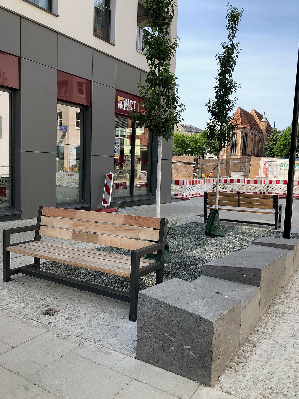 Zwei leere Holzbänke mit Metallgestell auf gepflastertem Platz vor einem Gebäude.