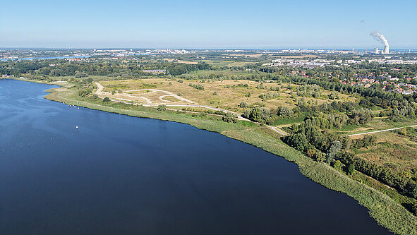 Luftaufnahme eines großen Gewässers mit angrenzender offener Landschaft und weiter Stadt