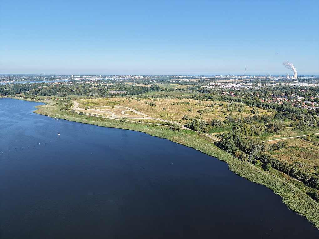 Luftaufnahme eines großen Gewässers mit angrenzender offener Landschaft und weiter Stadt