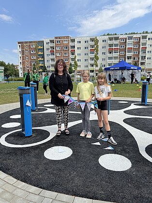 Drei Personen stehen auf einem großen Spielbrett im Freien vor Wohngebäuden, zwei Kinder und eine Erwachsene