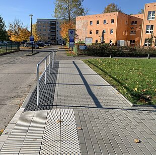 Gepflasterter Weg mit Metallgeländer zwischen Wohngebäuden und Grünflächen unter blauem Himmel
