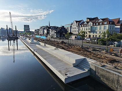 Uferpromenade mit moderner Sitzstufenanlage entlang eines ruhigen Stadthafens vor einer Häuserzeile unter blauem Himmel