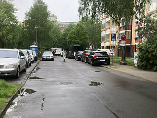 Blick auf eine regennasse Straße mit parkenden Autos beidseitig. Bäume und Gebäude im Hintergrund sind sichtbar.