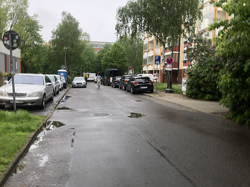 Blick auf eine regennasse Straße mit parkenden Autos beidseitig. Bäume und Gebäude im Hintergrund sind sichtbar.