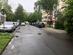 Blick auf eine regennasse Straße mit parkenden Autos beidseitig. Bäume und Gebäude im Hintergrund sind sichtbar.
