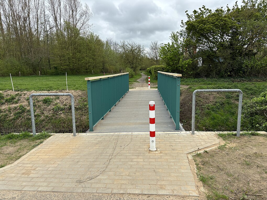 Grüner Fußgängerbrücke mit Metallgeländer über einem Wassergraben, davor ein Pfosten mit roten und weißen Streifen.