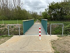 Grüner Fußgängerbrücke mit Metallgeländer über einem Wassergraben, davor ein Pfosten mit roten und weißen Streifen.