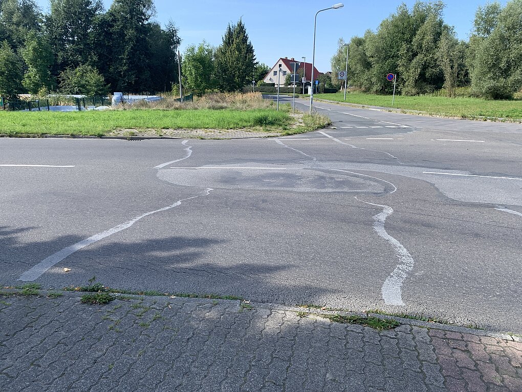 Straße mit mehreren großen Schlaglöchern und Rissen, umgeben von Bäumen und Häusern im Hintergrund