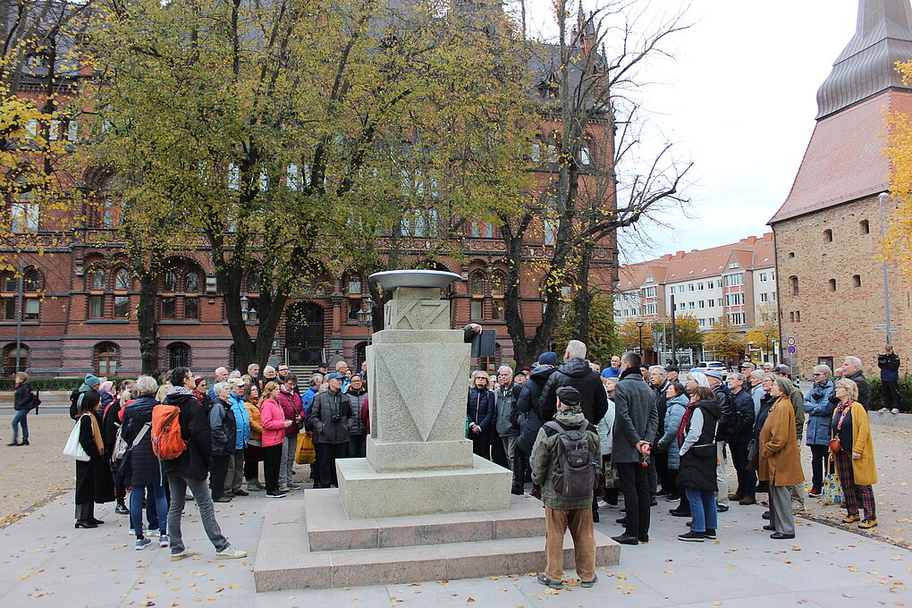 Menschenmenge vor einer Statue während einer Rede im Freien, Gebäude im Hintergrund