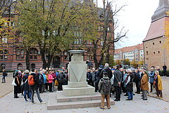 Menschenmenge vor einer Statue während einer Rede im Freien, Gebäude im Hintergrund