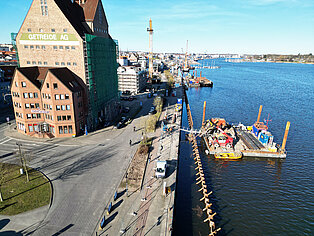 Blick auf den Stadthafen mit dem Gebäude 'Getreide AG', einer Baustelle am Wasser und der Warnow unter klarem Himmel