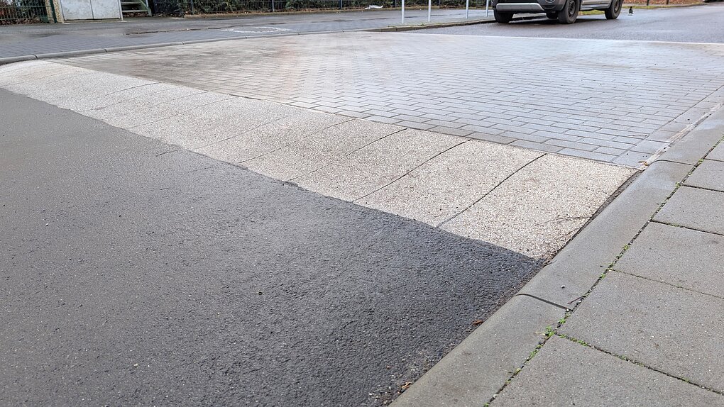Asphaltstraße mit unebenem Bereich und kleinen Steinen auf der Oberfläche.