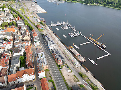 Luftaufnahme eines Stadthafens mit mehreren Segelbooten an Stegen, angrenzender Straße und Gebäuden mit roten Dächern