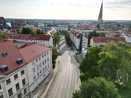 Luftaufnahme einer Stadt mit Straßen, Gebäuden und einem Kirchturm im Hintergrund.