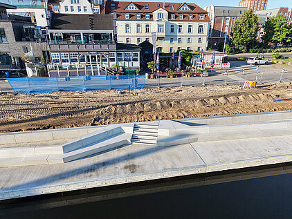 Uferpromenade mit breiter Treppe und Rampe am Wasser vor einer Baustelle und Gebäuden im Hintergrund