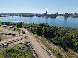 Luftaufnahme eines Flussufers mit Bauarbeiten an einem Weg und einer Stadt mit Kirchturm im Hintergrund