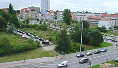 Straße mit mehreren Autos, daneben ein grüne Freifläche mit Bäumen und einem Weg, im Hintergrund Wohngebäude