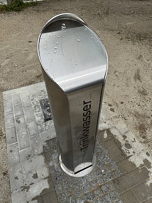 Metallsäule mit Aufschrift Trinkwasserbrunnen auf befestigten Boden im Freien.