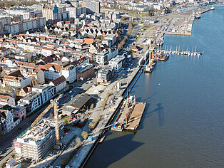 Luftaufnahme eines Stadthafens mit angrenzender Stadt, mehreren Gebäuden, Baustellen mit Kränen und Booten am Wasser