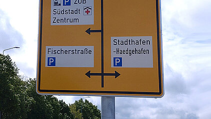 Gelbes Verkehrsschild mit Richtungspfeilen und Zielen wie Wismar, Warnemünde, HanseMesse, Zoo, ZOB, Südstadt, Zentrum, Fischerstraße, Stadthafen und Haedgehafen an einer Straße mit Autos und Kette am Straßenrand
