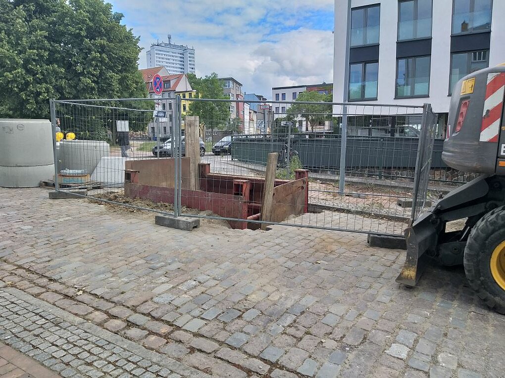 Blick auf eine eingezäunte kleine Baustelle mit Baugeräten im Boden..