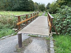 Holzbrücke mit Geländer über einen kleinen Bach in einem bewaldeten Gebiet.