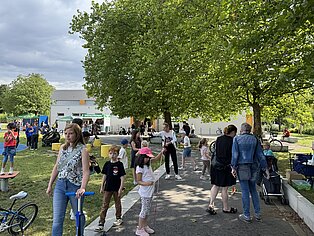 Menschen verschiedener Altersgruppen gehen und stehen unter Bäumen auf einem sonnigen Platz mit Gebäuden im Hintergrund