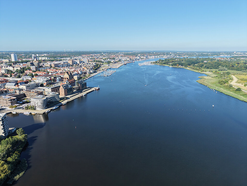 Luftaufnahme eines breiten Flusses mit Stadt und Hafen auf der linken Seite und grüner Landschaft rechts