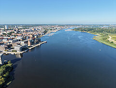 Luftaufnahme eines breiten Flusses mit Stadt und Hafen auf der linken Seite und grüner Landschaft rechts