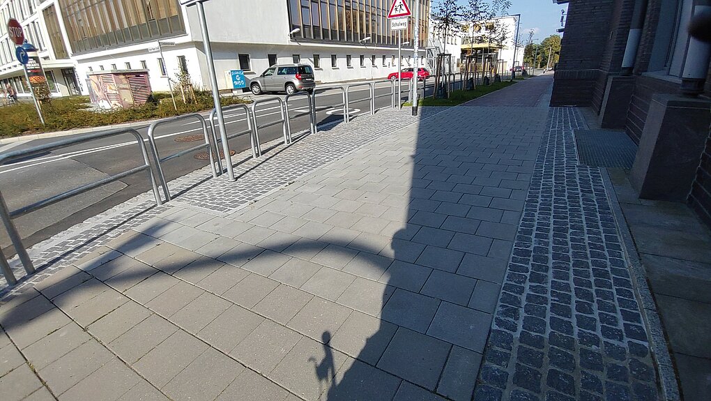 Straße mit Gehweg, Geländer und Gebäuden im Hintergrund bei sonnigem Wetter.