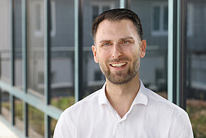 Junger MAnn mit kurzen Haaren und weißen Hemd lächelt in die Kamera. 