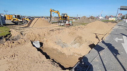 Baustelle mit großen Sandhaufen, Baggern und einer Baugrube neben einer Straße mit Rechtsabbiegerpfeil