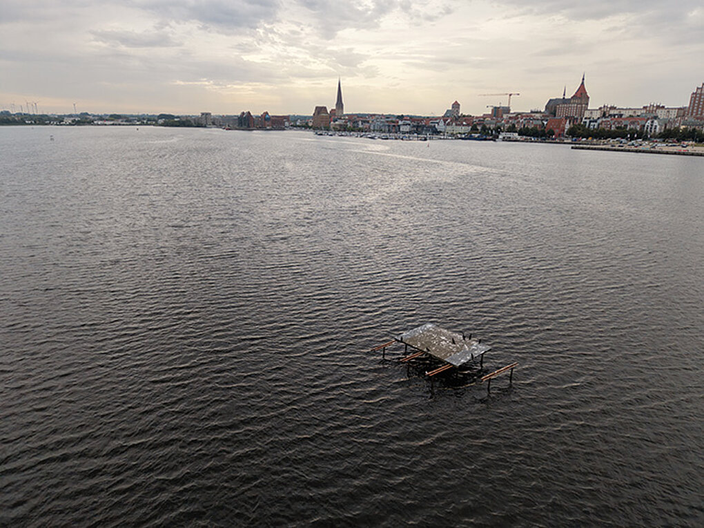 Blick über eine Wasserfläche mit einer kleinen defekten Plattform im Vordergrund und einer Stadt mit Kirchtürmen am Ufer im Hintergrund
