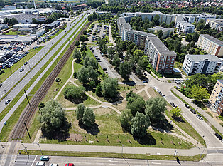 Luftaufnahme eines dreieckigen Parks mit Wegen und Bäumen neben Wohngebäuden und einer Straße mit Bahngleisen