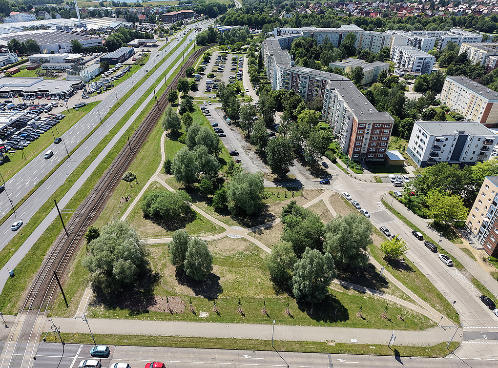 Luftaufnahme eines dreieckigen Parks mit Wegen und Bäumen neben Wohngebäuden und einer Straße mit Bahngleisen