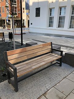Leere Holzbank mit Metallgestell auf gepflastertem Platz vor einem Gebäude mit weißen Wänden und Fenstern