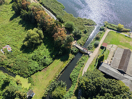 Luftaufnahme eines schmalen Wasserkanals mit Brücke, umgeben von Bäumen, Wiesen und Gebäuden neben einem größeren Gewässer