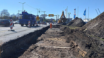 Baustelle an einer Straße mit aufgerissenem Boden, Bagger, Bauarbeiter in Warnkleidung und Fahrzeugen im Hintergrund