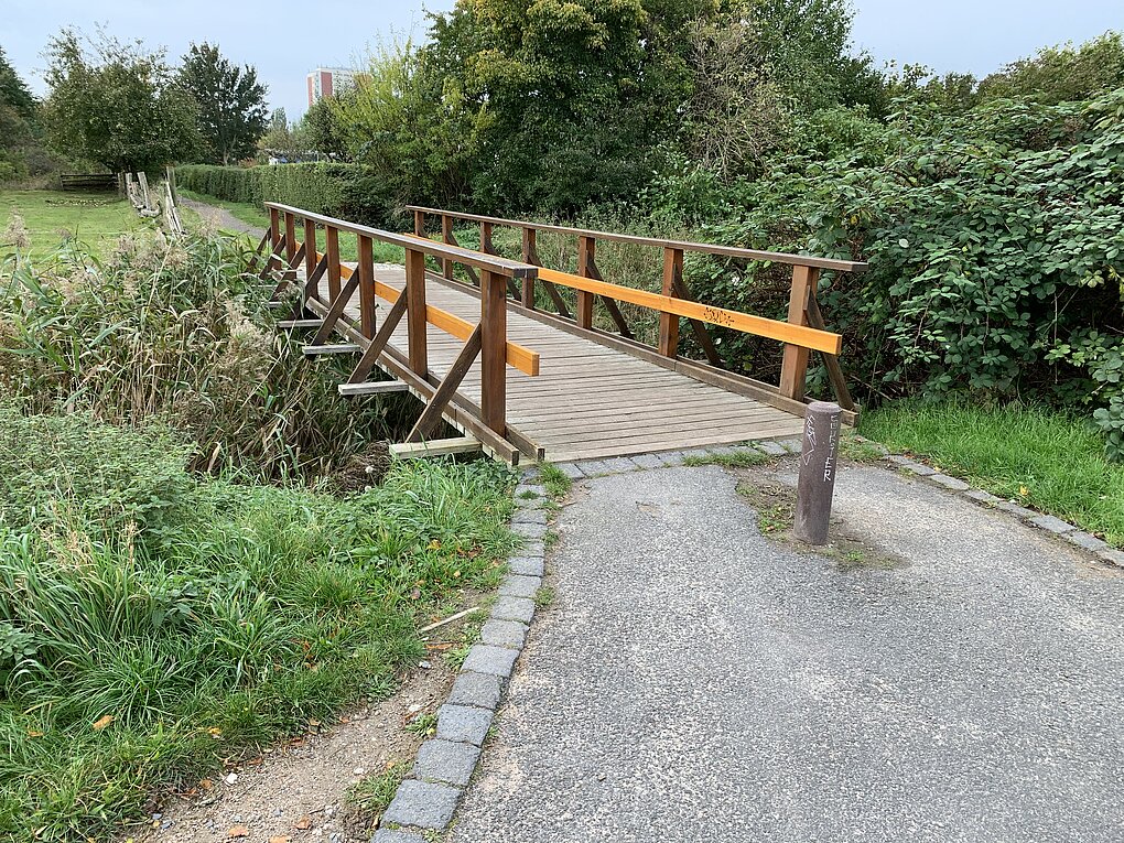 Holzbrücke über einen kleinen Wassergraben auf einem Kiesweg, umgeben von grünem Bäumen und Sträuchern.