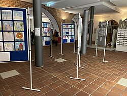Ausstellung mit mehreren Stellwänden, die Zeichnungen und Bilder zeigen, in einem Raum mit Kachelboden und großen Fenstern