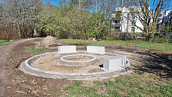 Kreisförmige Sitzanlage aus Betonblöcken auf einem Erdplatz in einem Park mit Bäumen und Wohngebäuden im Hintergrund